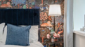 A bed with a dark headboard, a blue pillow, and a white pillow. There is a nightstand with a lamp and a vase of flowers. The wall behind the bed is decorated with a colorful mural.