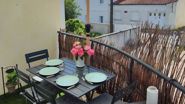 Un patio al aire libre con una mesa lista para cuatro personas, rodeada de sillas, un paraguas grande y un jarrón decorativo con flores.