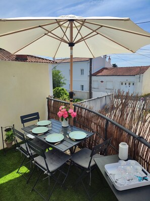 Une terrasse extérieure avec une table dressée pour quatre personnes, entourée de chaises, d’un grand parasol et d’un vase décoratif rempli de fleurs.