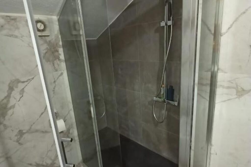 Un área de ducha con paredes de vidrio, suelo de mármol, un cabezal de ducha manual y un panel de control en la pared.
