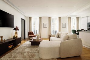 A modern living room with a sofa, a coffee table, a TV, and a large window with curtains. - Champs Elysees Fancy Home 2BDR 6PPL (Paris)