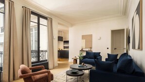 Ein Wohnzimmer mit einer Couch, einem Sessel, einem Couchtisch und Blick auf die Küche und den Balkon.