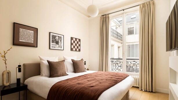 Ein Schlafzimmer mit Bett, Nachttisch mit Lampe, einem Fenster mit Vorhängen und Blick auf einen Balkon.