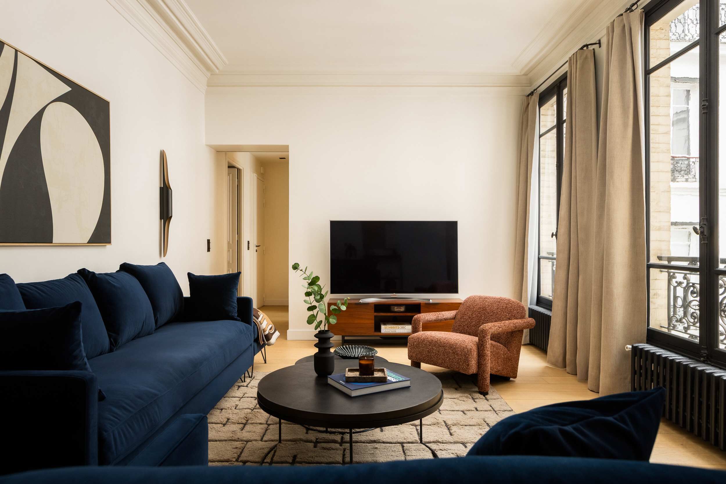 Uma sala de estar moderna com sofá, poltrona, mesa de centro, televisão e uma janela grande com cortinas.