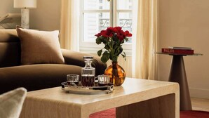 Camera d'albergo con divano, tavolino con bevande, vaso con fiori e vista attraverso una finestra con tende.