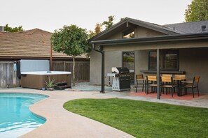 Un patio trasero con piscina, jacuzzi, barbacoa y un área de patio techada con sillas y mesas.