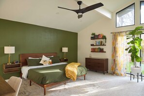 Un dormitorio con cama, mesitas de noche, cómoda, ventilador de techo, ventana con cortinas y una planta en maceta.