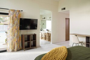 A modern bedroom with a television, a bed with a green cover, a desk with a chair, and a bathroom visible through an open door. - Retro Ranch | Home Theater, Pool/Spa, Pet Friendly (Fresno)