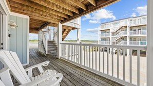 Una terraza de madera con barandas blancas, una escalera que conduce a un nivel superior y vista de un edificio frente a la playa.