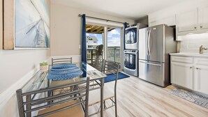 Una cocina moderna con un refrigerador de acero inoxidable, una mesa de comedor de vidrio y vista a una casa a través de una puerta abierta.