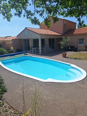 A swimming pool with a white edge, a covered patio area with tables and chairs, and a building with a tiled roof in the background. - Villa Océane with heated swimming pool. (Les Sables-d'Olonne)