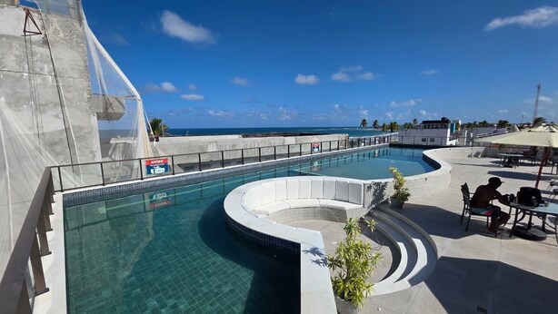 A pool area with a curved design, a sign indicating AVISO, and a view of the ocean. - Ora Beach poucos passos do mar by LP (Ipojuca)