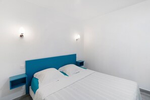 Una cama con cabecera azul, ropa de cama blanca y dos mesitas de noche con lámparas de pared.