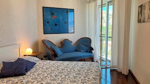 Ein Schlafzimmer mit einem Bett, einem blauen Sessel, einem Wandbild und einem Fenster mit Vorhängen.