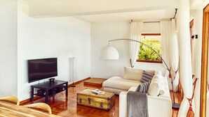 A modern living room with a flat-screen TV, a wooden dining table, a sofa with cushions, a floor lamp, and a window with curtains. - Wonderfull view! (Èze)