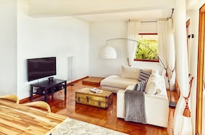 A modern living room with a flat-screen TV, a wooden dining table, a sofa with cushions, a floor lamp, and a window with curtains. - Wonderfull view! (Èze)