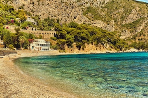 Ett hotell vid kusten med en bergig sluttning, kristallklart blått vatten och en strand med småsten.