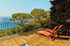 A wooden deck with two lounge chairs, a pool, and a view of the sea and trees.
