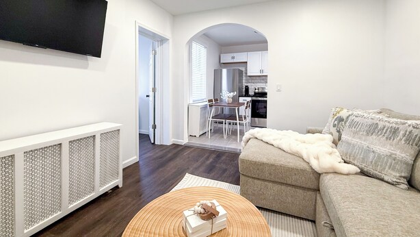 Una sala de estar con un sofá, una mesa de centro con una caja, un televisor montado en la pared, y una cocina visible a través de un arco.