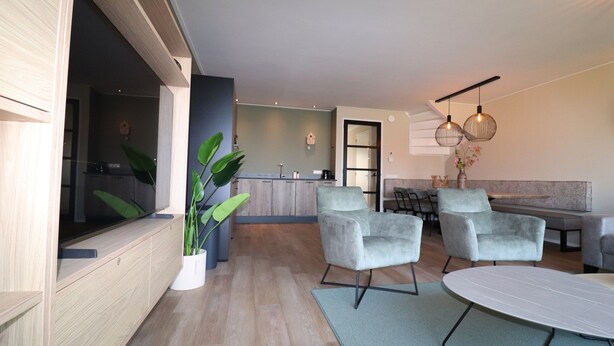 Een moderne woonkamer met een bank, twee fauteuils, een salontafel en een televisie. Achtergrond toont een keuken met een eethoek.