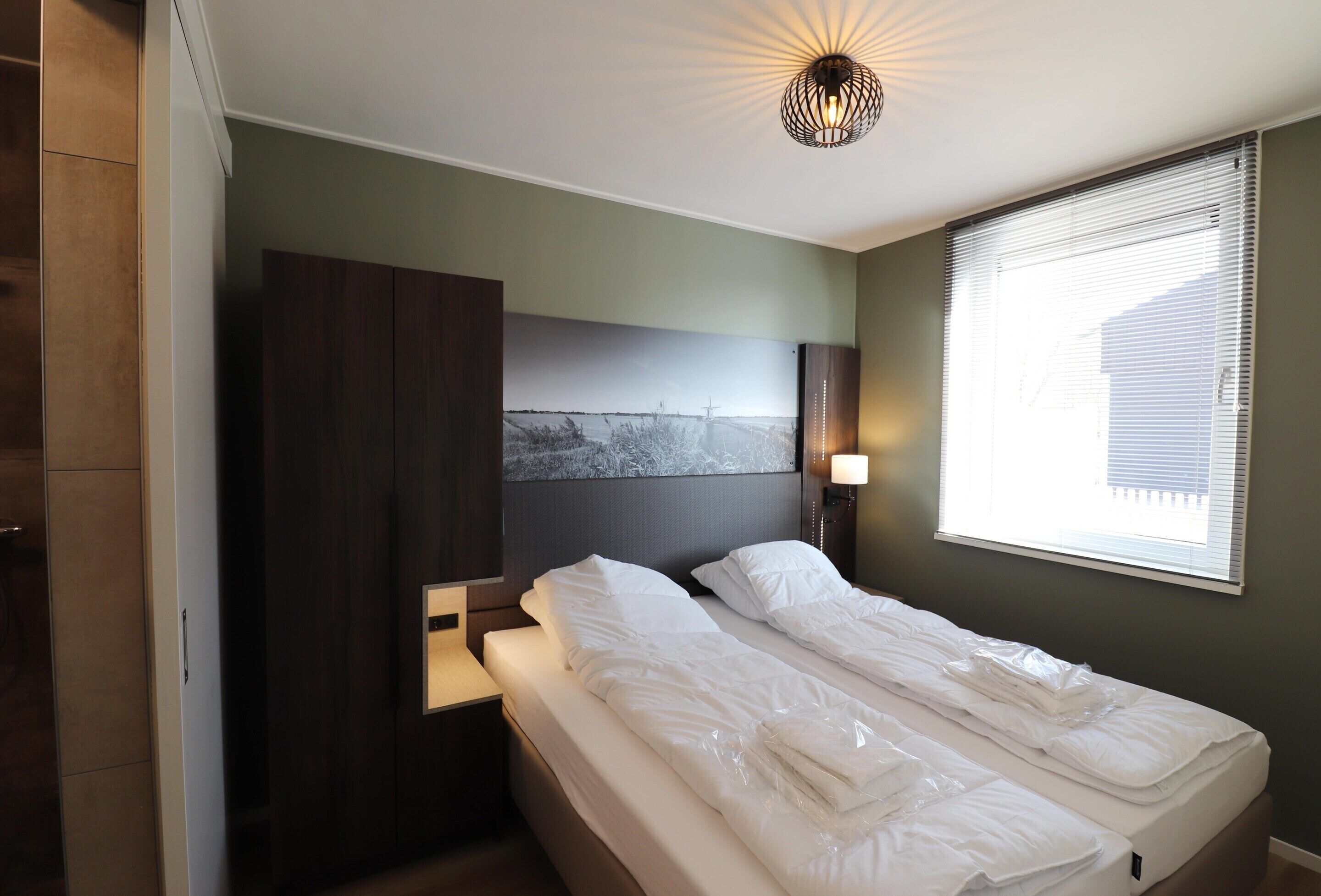 A bedroom with two beds, a nightstand, a wall-mounted lamp, and a window with blinds.
