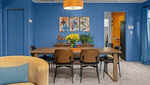 A dining area with a blue wall, a wooden table, and six chairs. There are two paintings on the wall, a vase with flowers, and a hanging light fixture. - Black Diamond | Mid-Century Modern Hot Tub Retreat (Clallam County)