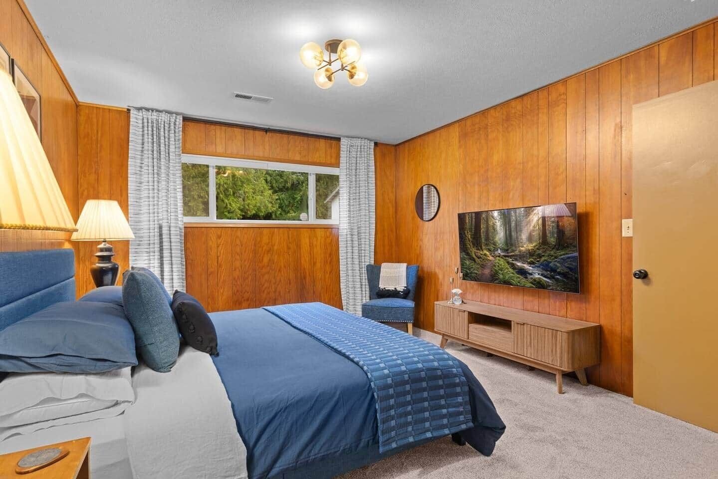 A bedroom with a bed, a TV, a window, and a wooden wall.