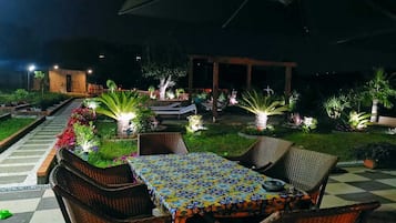 Un área de comedor al aire libre con mobiliario de mimbre y un mantel estampado, rodeada de plantas iluminadas y un sendero que conduce a un edificio.
