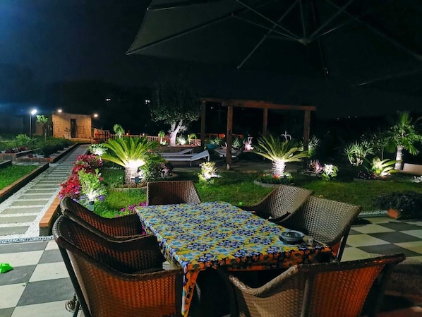 An outdoor dining area with wicker furniture and a patterned tablecloth, surrounded by illuminated plants and a pathway leading to a building.