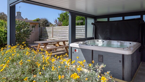 Un bain à remous installé sous une terrasse couverte, avec une table et des chaises en bois, le tout entouré de fleurs jaunes.