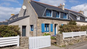 Une maison en pierre avec un toit en pente, de nombreuses fenêtres dotées de volets bleus et une clôture en bois peinte en blanc.