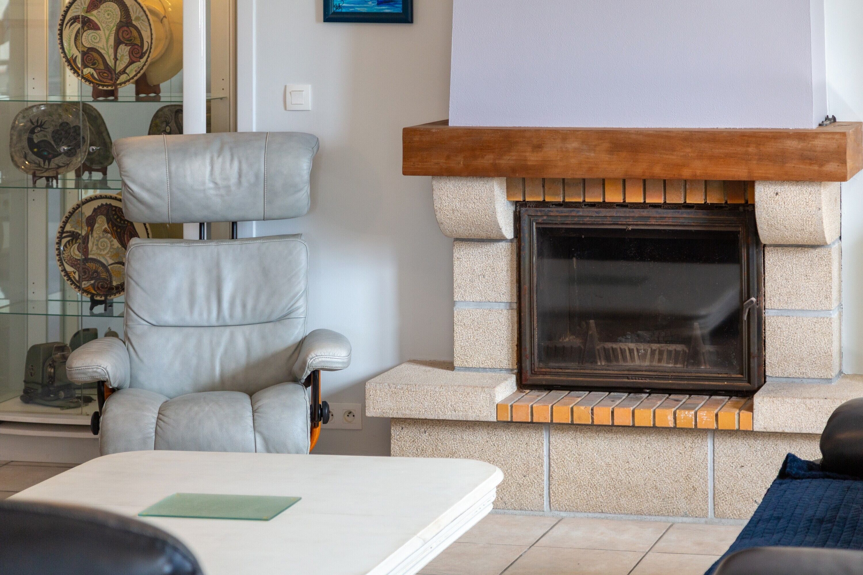 A living room with a fireplace, a leather armchair, and a glass display case with decorative plates.