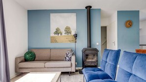 A living room with a sofa, a blue couch, a table, a fireplace, and a painting on the wall. - 5-guest Forest Bungalow in Ewijk (Ewijk)