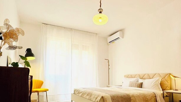 Quarto com cama, mesa de cabeceira, abajur e janela com cortinas.