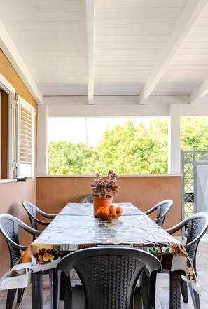 A covered outdoor dining area with a table set for two, surrounded by chairs, a potted plant, and a window with a view of greenery. - Ruralholidays Experience in the Heart of Salento (Castri di Lecce)