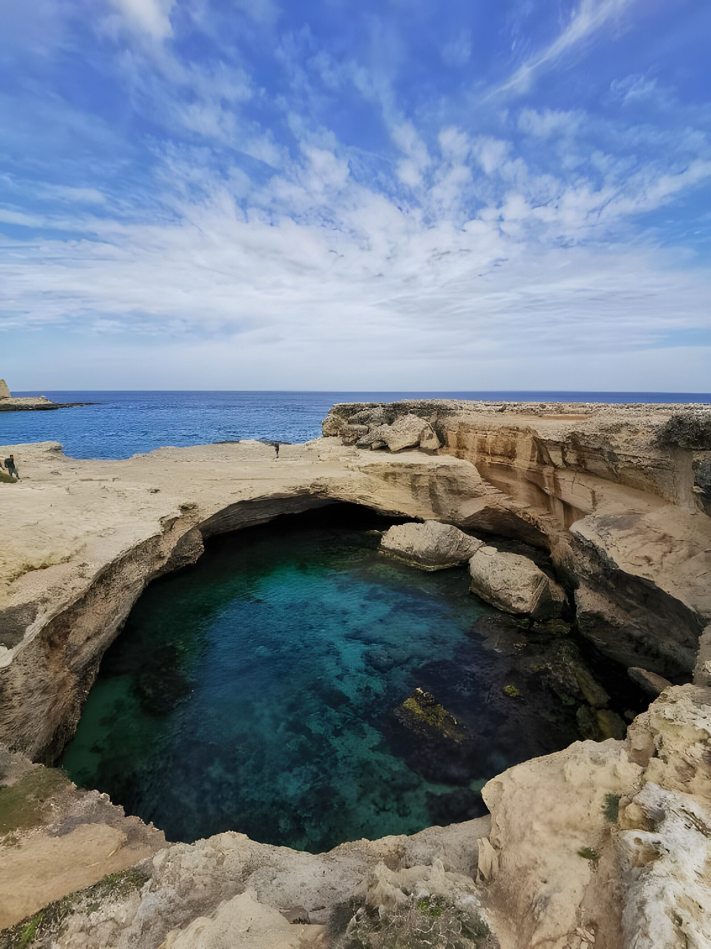 Une formation rocheuse naturelle avec une étendue d’eau claire, une rive rocheuse et une vue lointaine sur la mer, le tout sous un ciel partiellement nuageux.