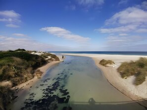 Una playa con un canal de agua cristalina, orillas arenosas y vegetación escasa.