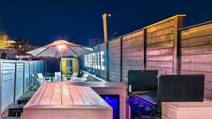 An outdoor patio area with a wooden table, chairs, a grill, and a neon sign. - Walk to Broadway • 14 Beds • Pool/Hot Tub • Sauna! (Nashville)