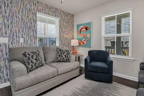 Un salon avec un canapé gris, des coussins à motifs, un fauteuil bleu, une petite table avec une lampe et un tableau représentant des carpes koï au mur.