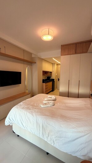 Ein Schlafzimmer mit einem Bett, einem an der Wand montierten Fernseher, einem Spiegel und einer kleinen Küchenzeile im Hintergrund.