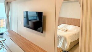 Ein modernes Hotelzimmer mit einem an der Wand montierten Flachbildfernseher, einer Holzbank, einem Bett mit weißer Matratze und einem großen Spiegel.