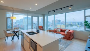 A modern kitchen with a central island, dining area with chairs, a sofa, and large windows offering city views. - West Coast City Living (Victoria)