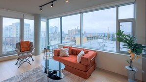A modern living room with a large window, a sofa, a chair, and a small table. - West Coast City Living (Victoria)
