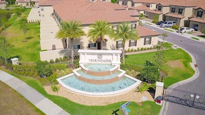 A residential area with a central building labeled 'The Fountains,' surrounded by single-family homes with red-tiled roofs, a landscaped area with palm trees, and a gated entrance. - Gorgeous Snowbird-friendly Home With Pool, hot Tub, & gym - Close to Theme Parks (Davenport)