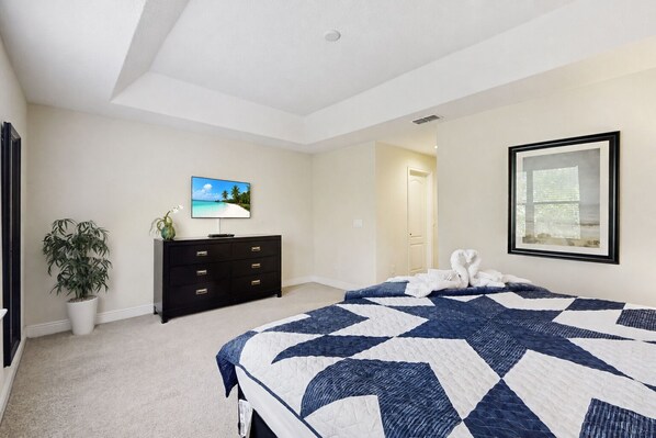 Un dormitorio con cama, cómoda, televisor, planta y un cuadro en la pared.