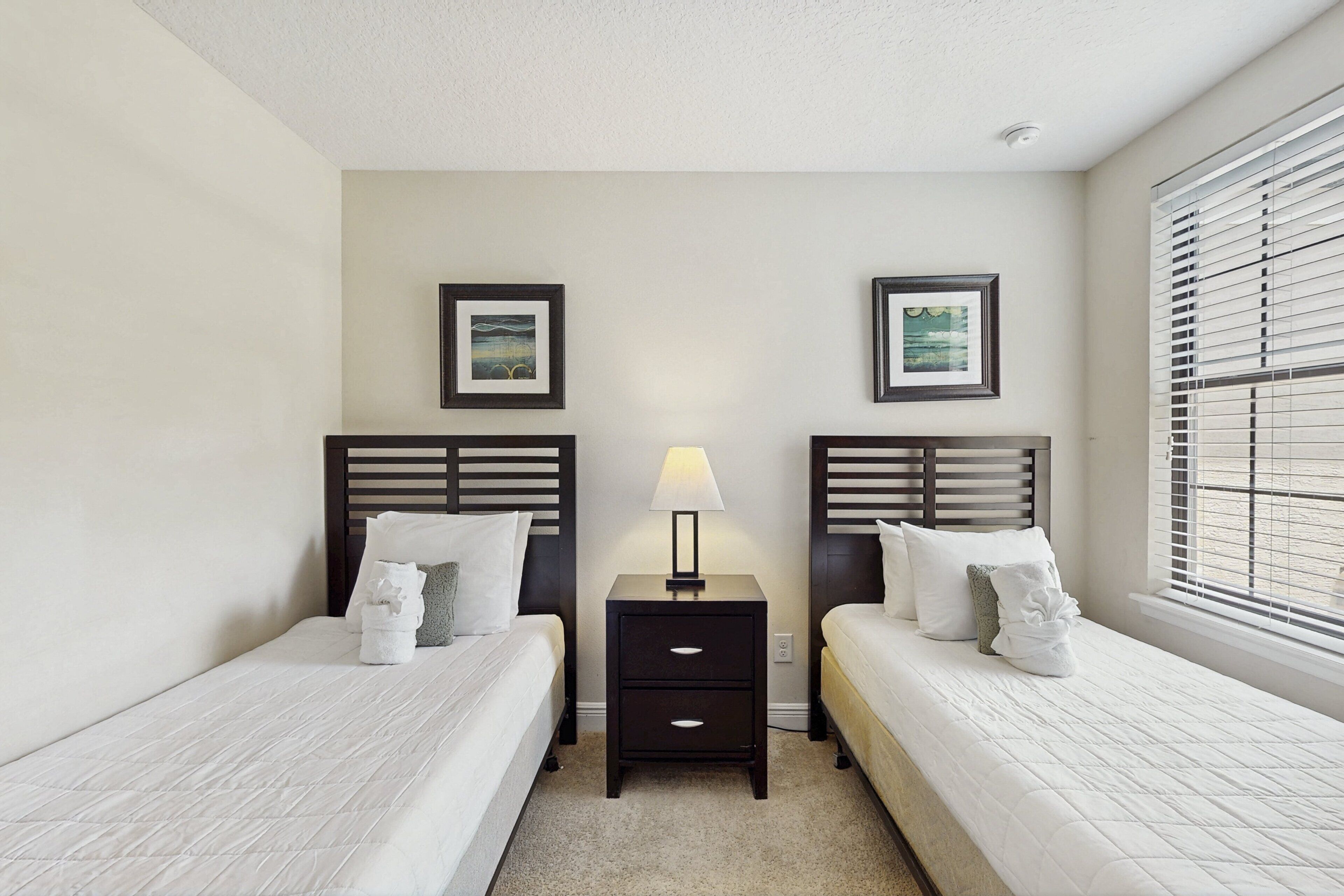 A hotel room with two beds, a nightstand with a lamp, and two framed pictures on the wall.