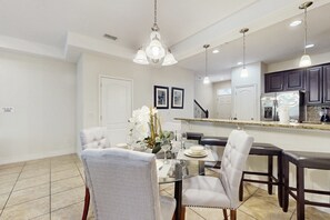 A dining area with a glass table, grey upholstered chairs, a chandelier, and a kitchen with dark cabinets and a stainless steel refrigerator. - Gorgeous Snowbird-friendly Home With Pool, hot Tub, & gym - Close to Theme Parks (Davenport)