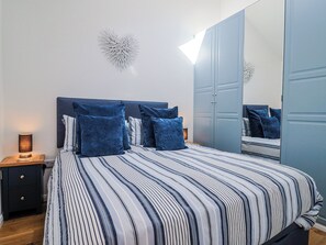A bedroom with a bed, a nightstand, a lamp, a mirror, and a wardrobe. - Le Petit Glen (Lyme Regis)