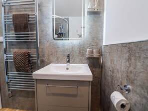 A bathroom with a white sink, a towel rack with a brown towel, a mirror, and a toilet paper holder. - Le Petit Glen (Lyme Regis)