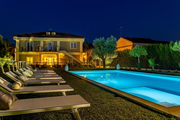 Uma piscina com espreguiçadeiras, uma casa com varanda e um jardim bem iluminado à noite.
